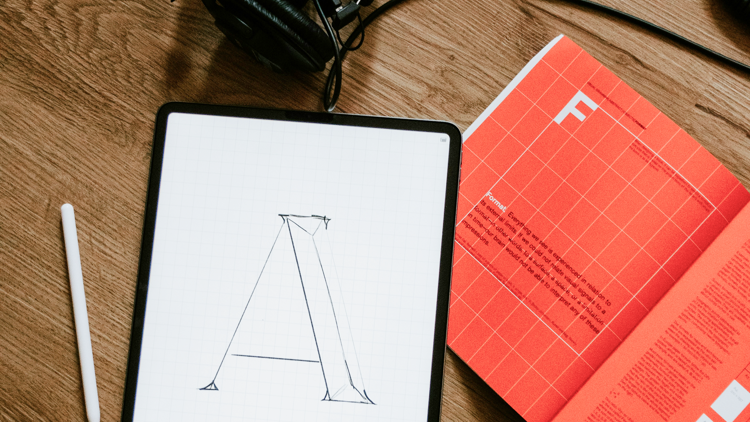 Surfplatta med skiss, hörlurar och bok på golv.