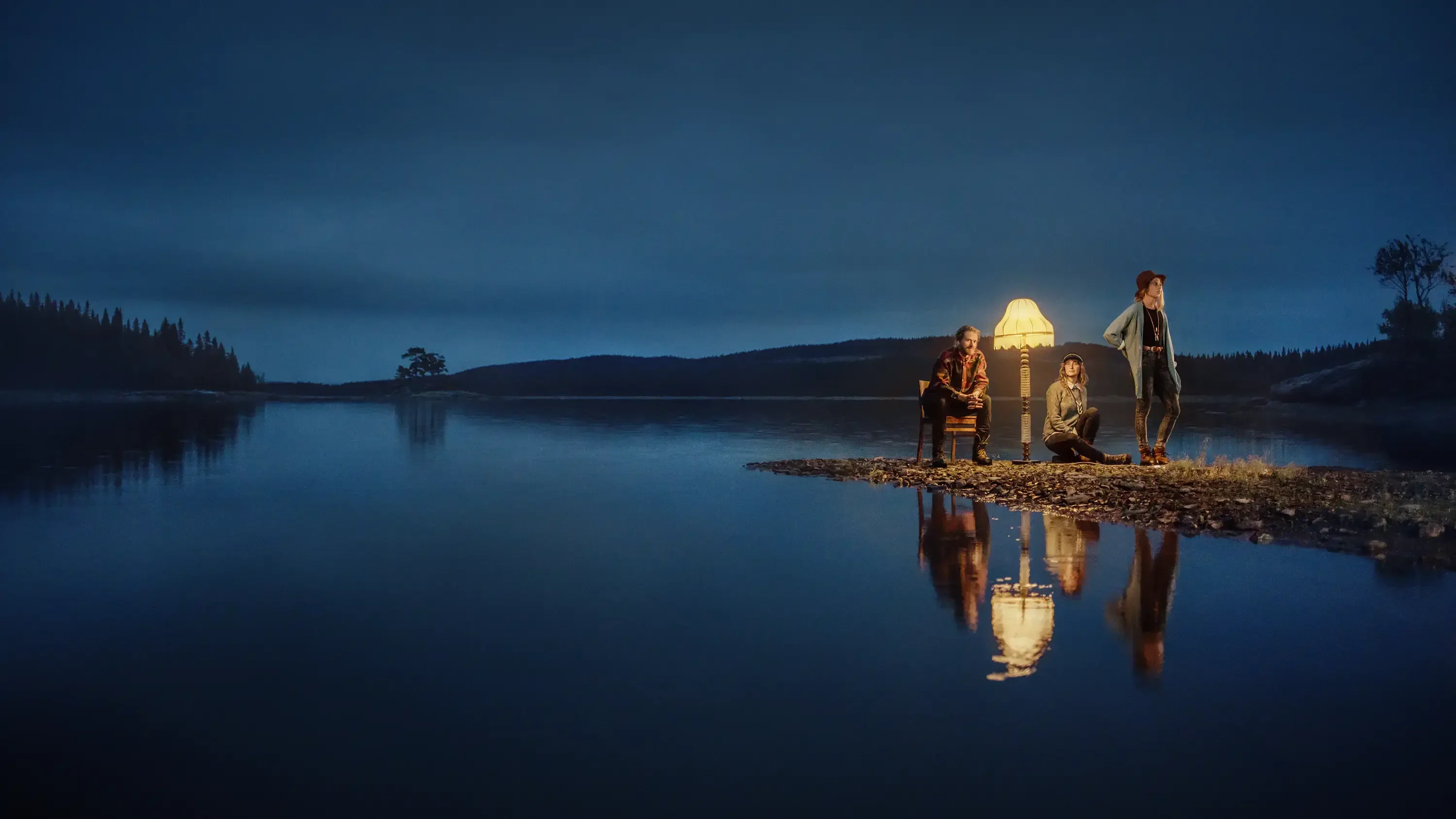 three persons standing by a lake with a lit lamp when it's turning dark
